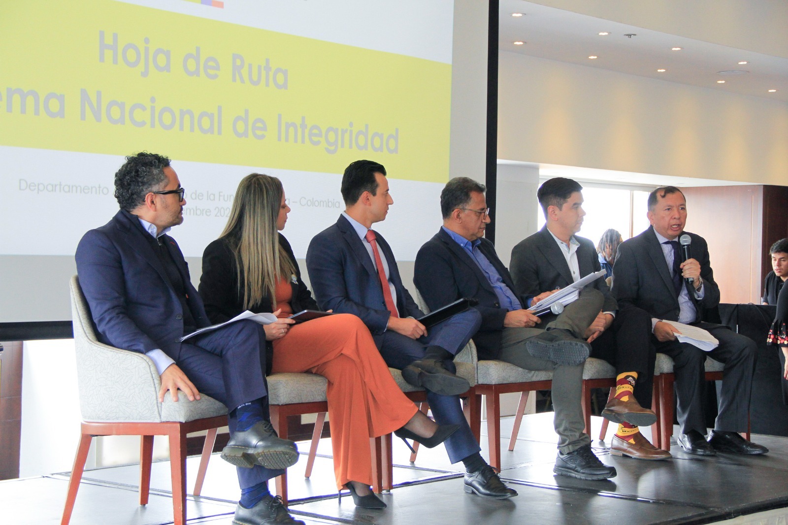 Cinco hombres y una mujer sentados en sillas sobre una tarima 