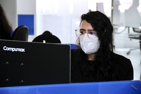 Mujer sentada en un puesto de trabajo, con tapabocas y gafas, mirando la pantalla de un computador