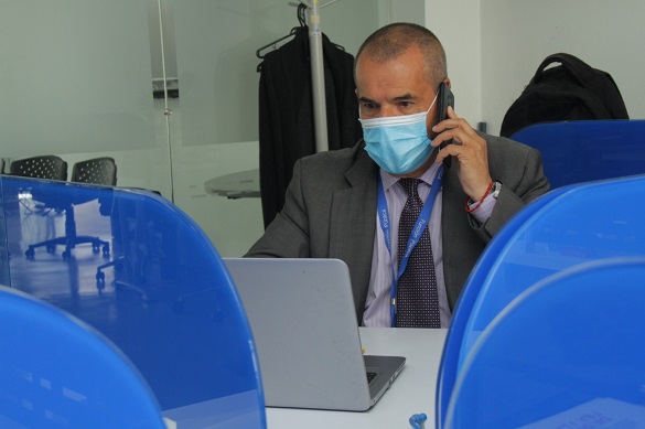 Hombre con tapabocas hablando por teléfono celular y trabajando en un computador, en un puesto o estación de trabajo