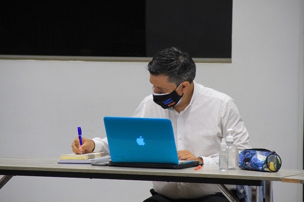 Hombre con tapabocas sentado frente a una mesa en donde hay un computador portátil y con una mano está escribiendo sobre una libreta ubicada a un lado del computador