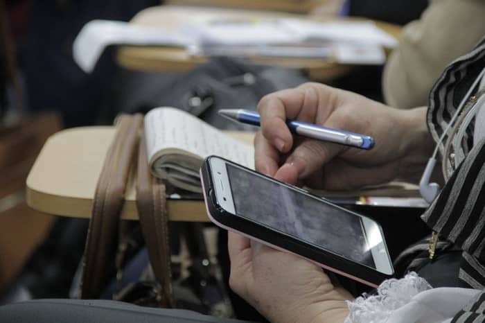 Se ven las manos de una persona que está sentada en un pupitre sosteniendo un teléfono celular
