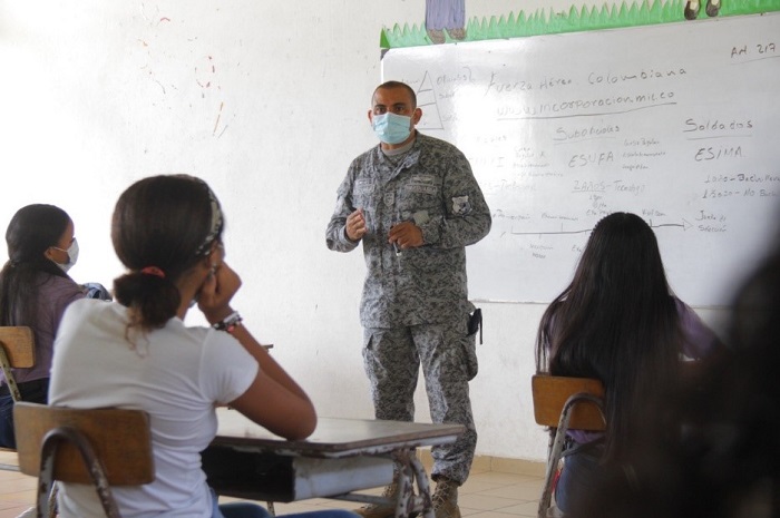 Militar con tapabocas dando instrucciones en un salón de clases frente a un grupo de mujeres sentadas en pupitres