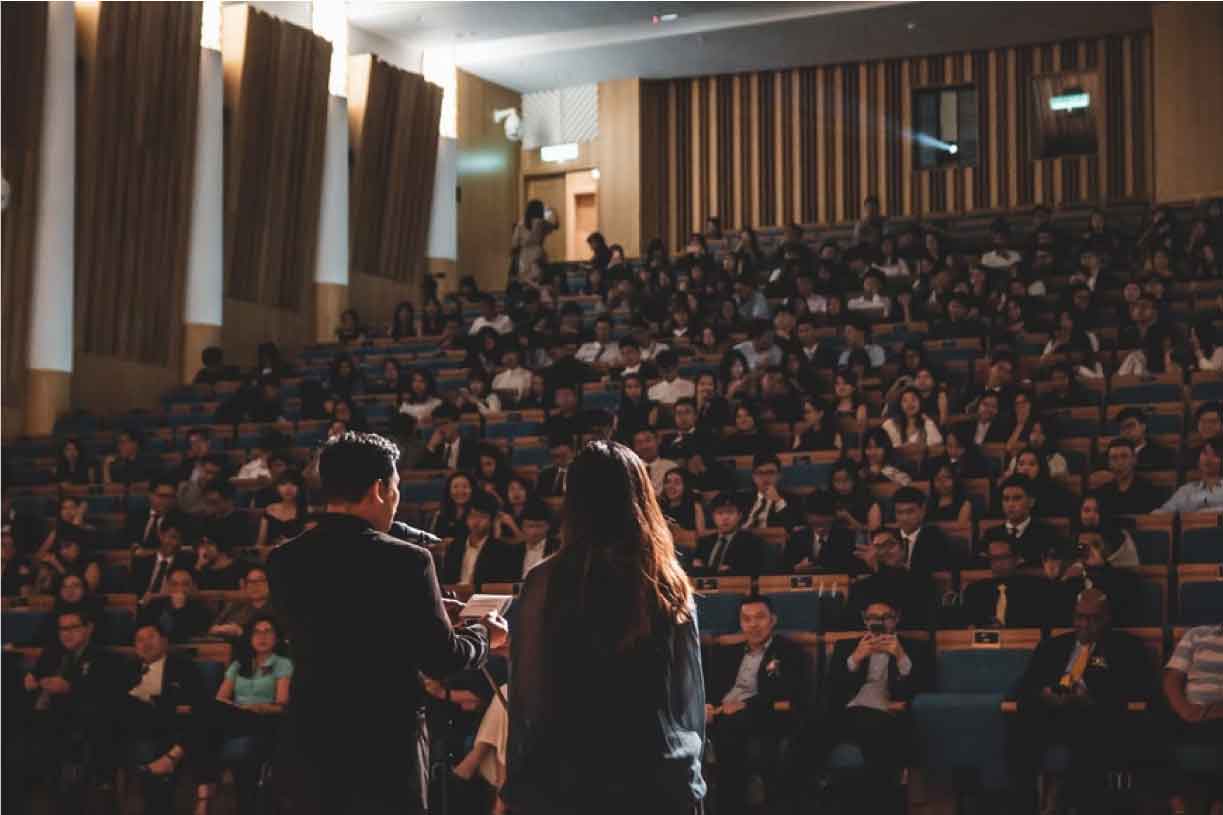 Dos personas hablando frente a un auditorio lleno de personas